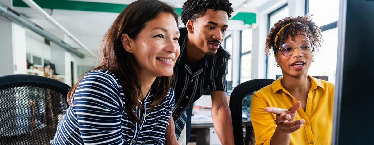 Three smiling business people are collaborating on a project, gathered around a desktop computer in a bright, modern office.