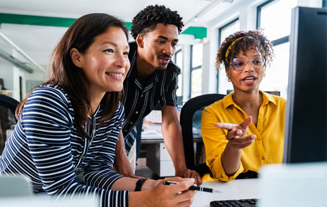 Three smiling business people are collaborating on a project, gathered around a desktop computer in a bright, modern office.